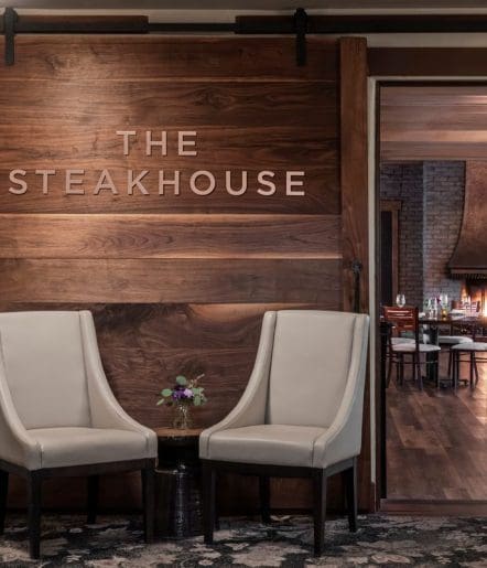 Two beige armchairs with a small round table and a floral arrangement sit in front of a wooden wall with THE STEAKHOUSE sign; among Paso Robles Restaurants, its dining room with set tables is visible through an open doorway.