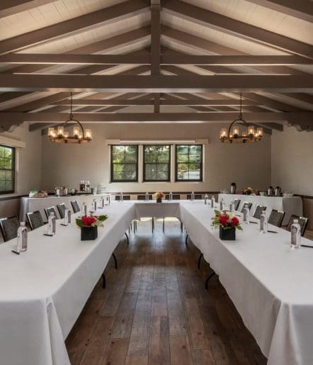 A well-lit meeting room with wooden floors and exposed beams, featuring two U-shaped tables covered with white cloths, each place set with a water bottle and notepad. Flower arrangements decorate the tables.