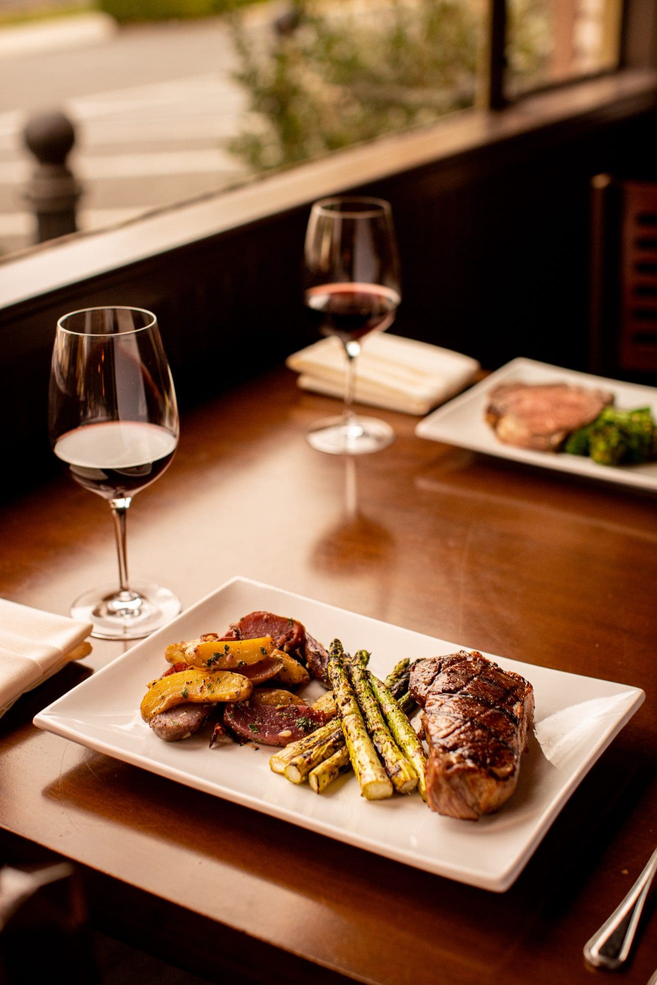 A neatly set Steakhouse Paso Robles table features a steak, roasted potatoes, and grilled asparagus on a white plate, accompanied by two glasses of red wine and another plate with greens and meat in the background.
