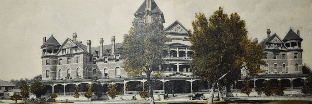 Historic black-and-white photo of a large Victorian-style building with turrets, multiple chimneys, a tall central tower, and a wraparound porch, with trees and a vintage car in front.