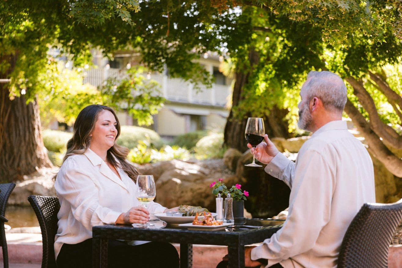 A woman and a man sit at an outdoor table under trees at a Steakhouse Paso Robles, enjoying a meal and toasting with glasses of wine. Sunlight filters through the branches as a small flower pot decorates the table.