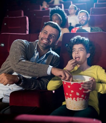 A man and a child sit together in a movie theater at one of the Paso Robles Events, sharing a large tub of popcorn and smiling. Other people are seated behind them, also watching the movie. The theater seats are red.