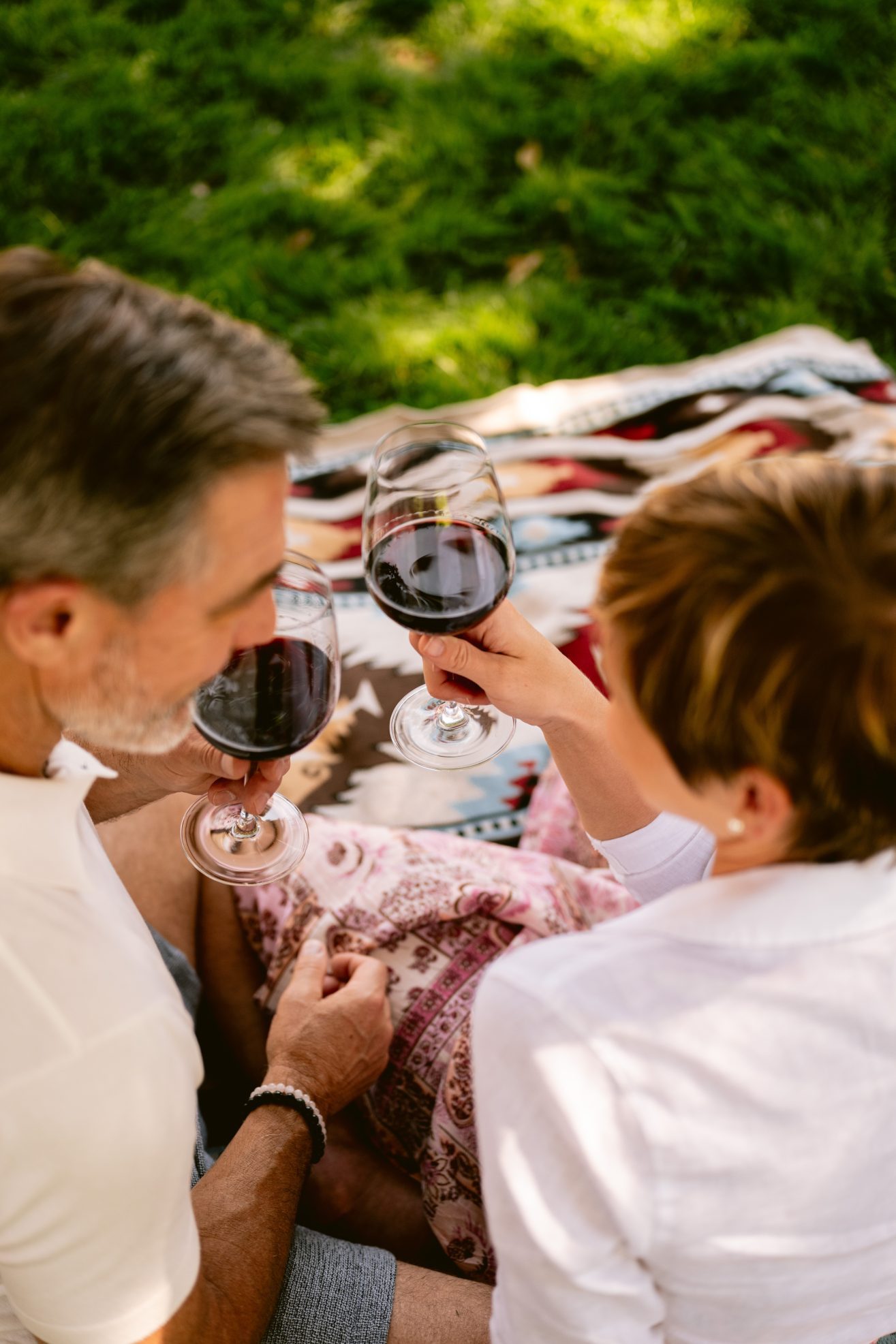 A Man and a woman sitting on a picnic blanket drinking red wine together.
