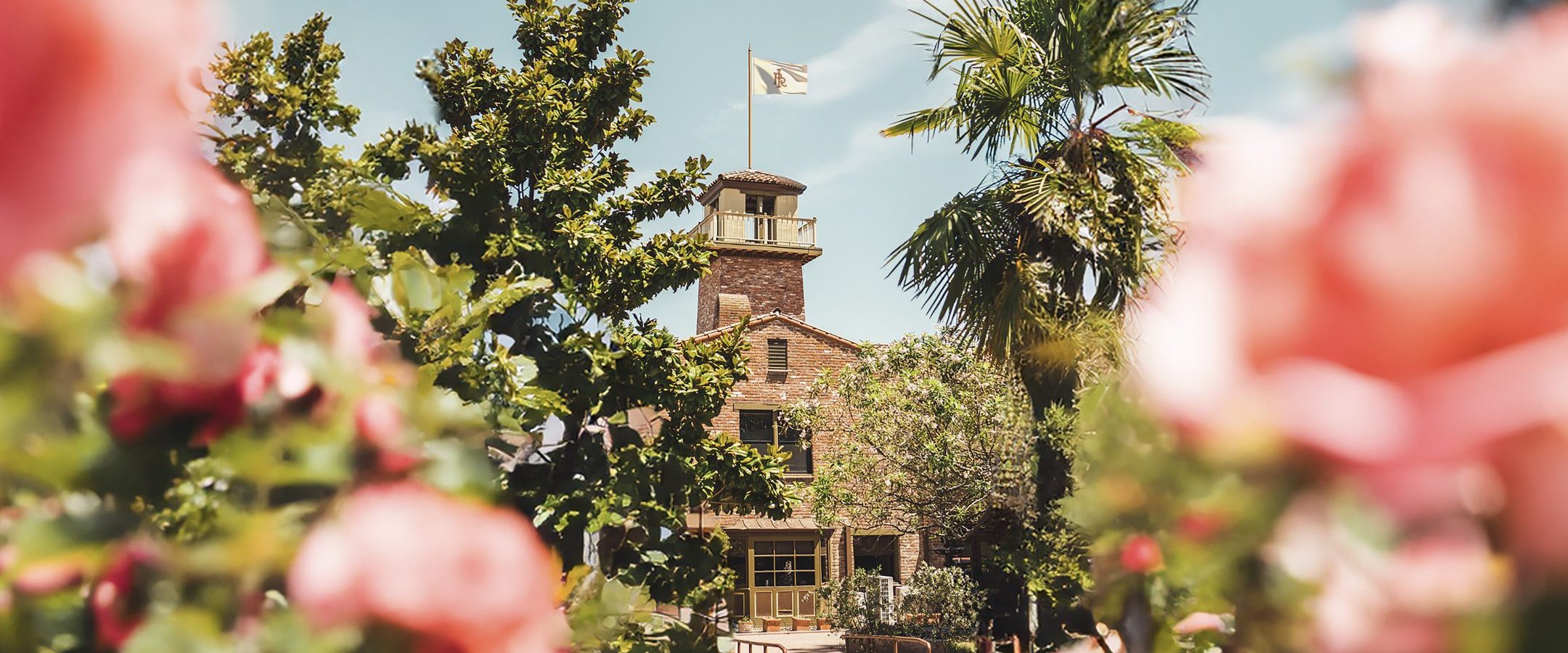 A brick building with a tower and flag stands in the sunlight, surrounded by palm trees and blooming pink flowers in the foreground. The sky is blue with wispy clouds.