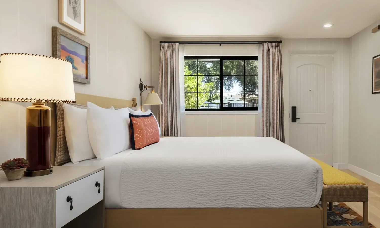 A cozy bedroom with a neatly made bed, white bedding, decorative pillows, bedside table with a lamp and small plant, wall art, a window with trees outside, and a door to the right.