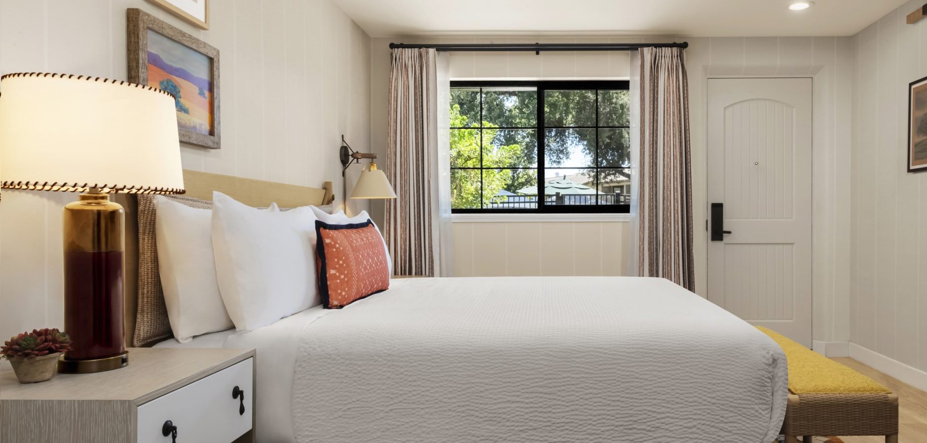 A cozy bedroom with a neatly made bed, white bedding, decorative pillows, bedside table with a lamp and small plant, wall art, a window with trees outside, and a door to the right.