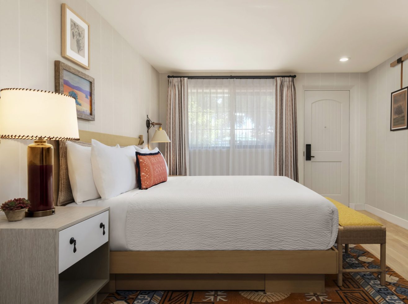 A cozy, neatly made bed with white linens and decorative pillows sits next to a nightstand with a lamp and a small plant in a bright, modern bedroom with wall art and a window letting in natural light.