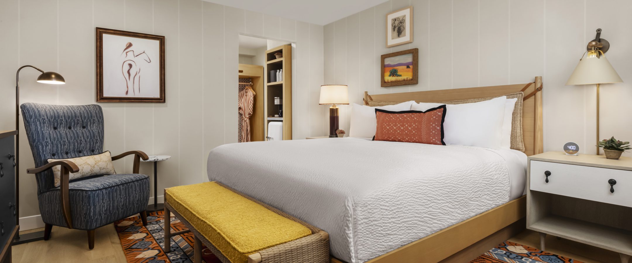 A cozy, modern bedroom with a large bed, white bedding, yellow bench, blue armchair, nightstands with lamps, wall art, and a colorful patterned rug on light wood flooring.