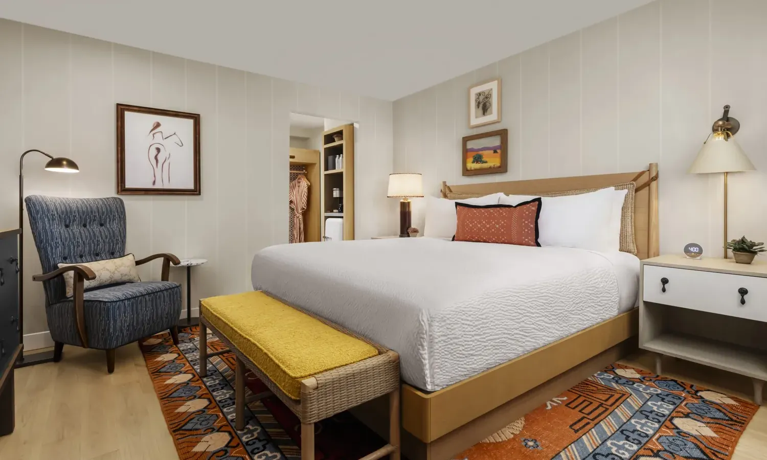 A cozy, modern bedroom with a large bed, white bedding, yellow bench, blue armchair, nightstands with lamps, wall art, and a colorful patterned rug on light wood flooring.