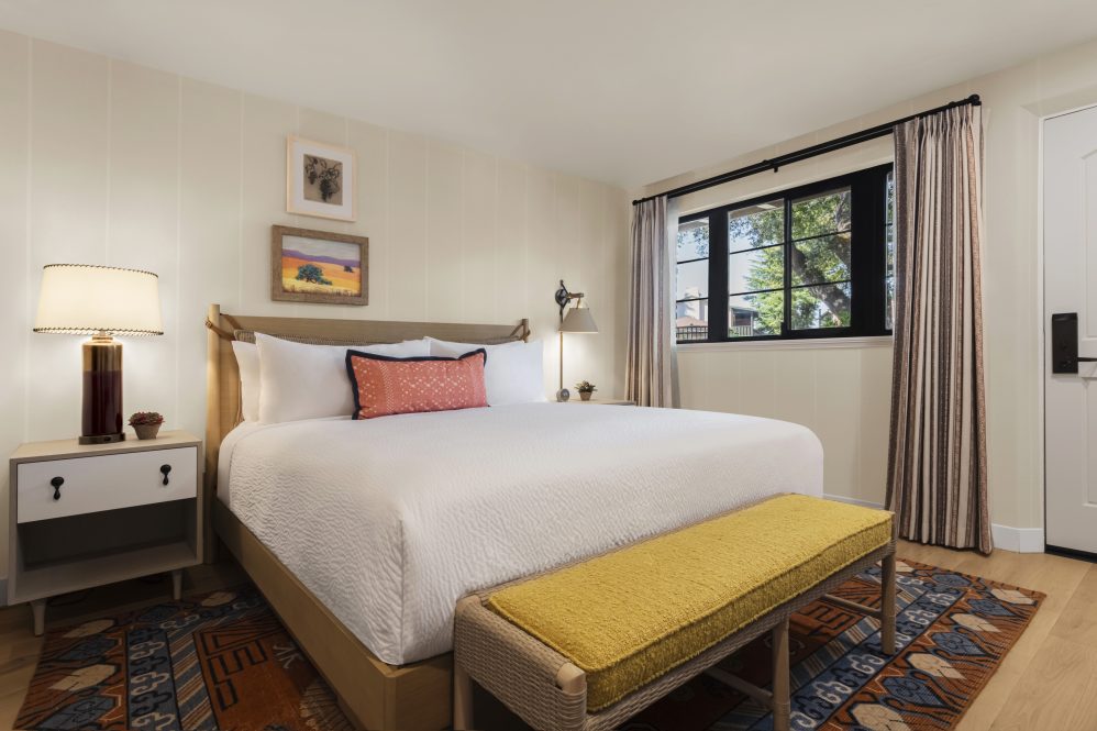 A cozy bedroom with a large neatly made bed, a yellow bench at the foot, two nightstands with lamps, framed art on the wall, a window with curtains, and a patterned rug on the floor.