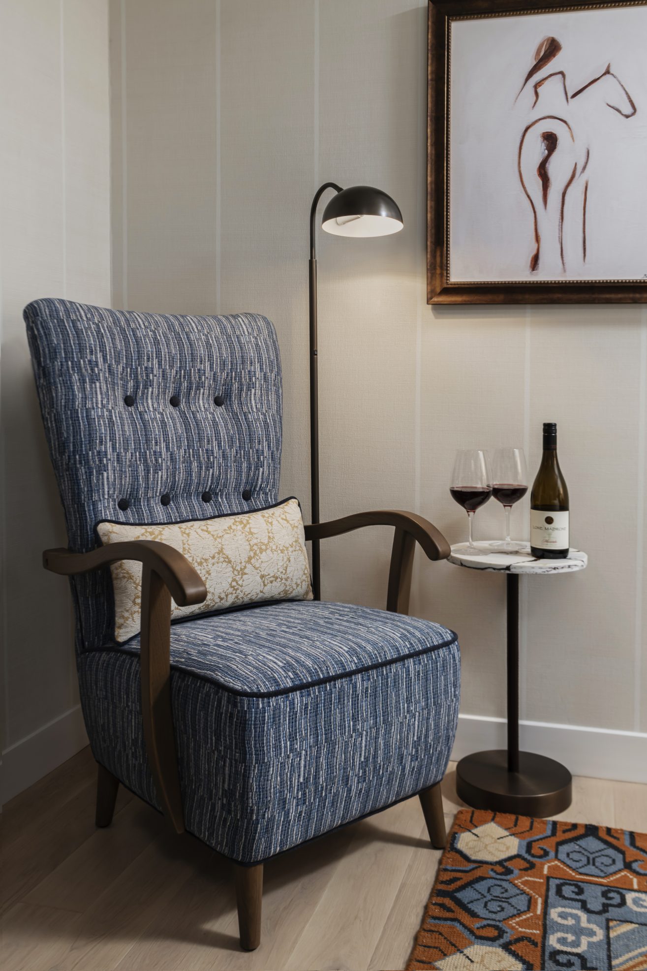 A blue patterned armchair with a decorative pillow sits beside a small round table holding two glasses of red wine and a wine bottle. A floor lamp and abstract horse artwork are in the background.