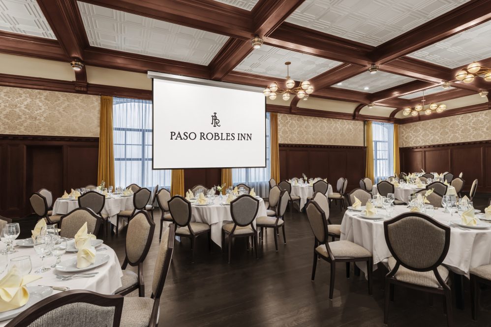 Elegant banquet hall with round tables set for dining, beige upholstered chairs, folded napkins, and glassware. A large projector screen at the front displays “Paso Robles Inn.” Chandeliers hang from a coffered ceiling.