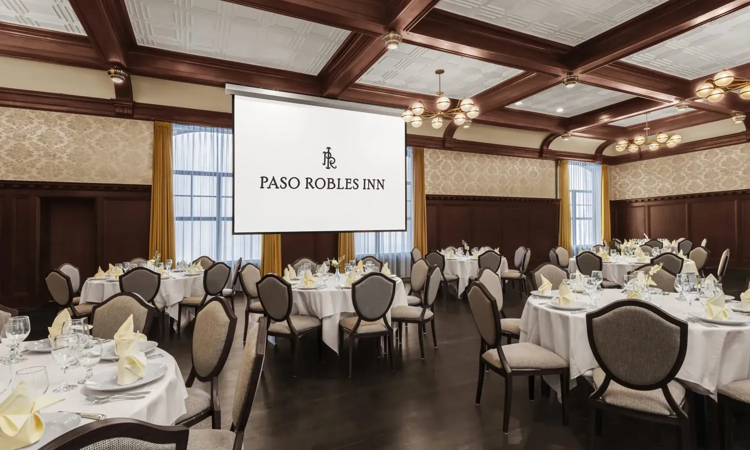 Elegant banquet hall with round tables set for dining, beige upholstered chairs, folded napkins, and glassware. A large projector screen at the front displays “Paso Robles Inn.” Chandeliers hang from a coffered ceiling.