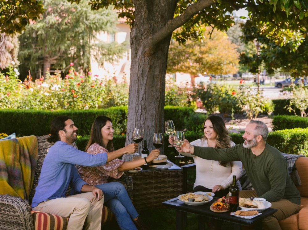 Four people sit at an outdoor table under a tree, smiling and raising wine glasses together. Food and a bottle of wine are on the table, and the background is a garden with green hedges and flowering plants.