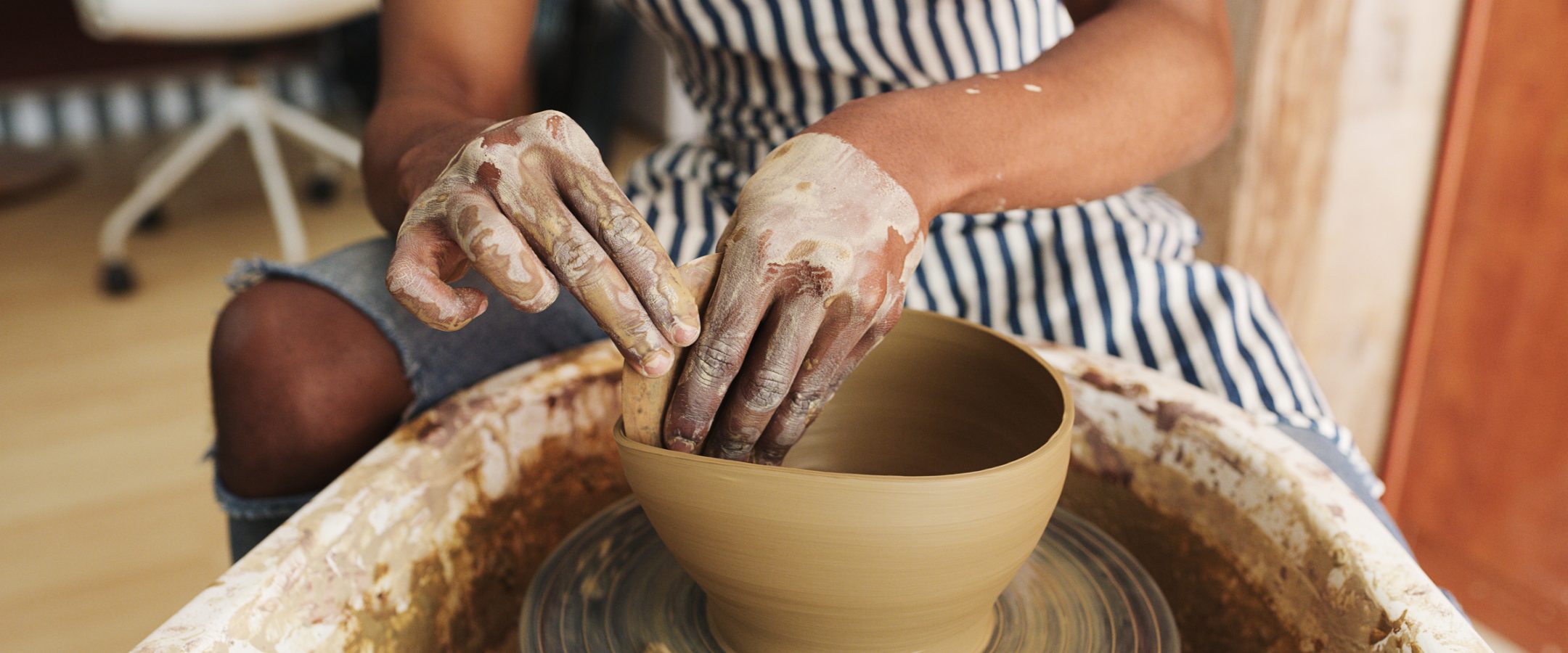 A person wearing a striped apron shapes a clay bowl on a pottery wheel, with their hands covered in wet clay. The surrounding area appears to be a pottery studio.