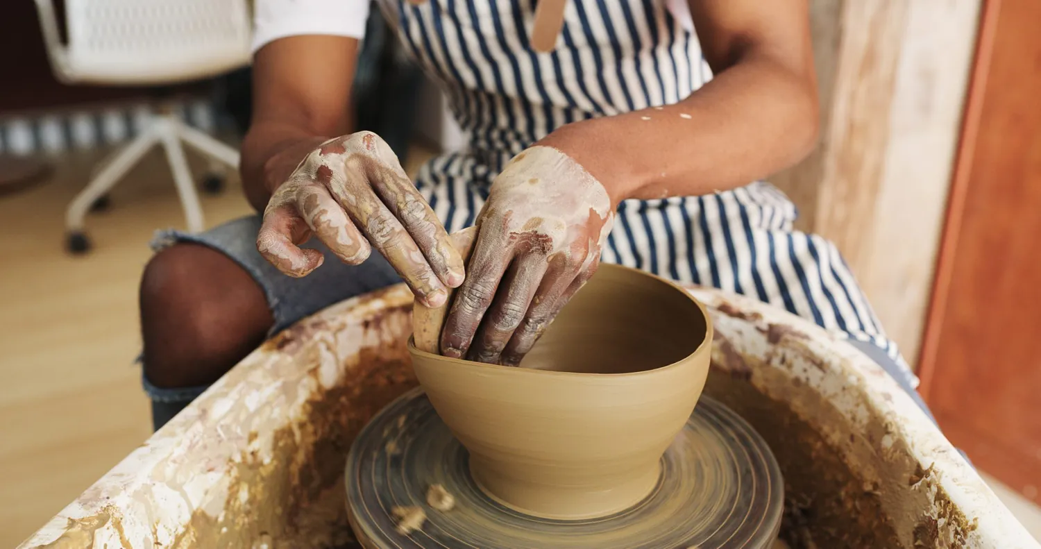 A person wearing a striped apron shapes a clay bowl on a pottery wheel, with their hands covered in wet clay. The surrounding area appears to be a pottery studio.