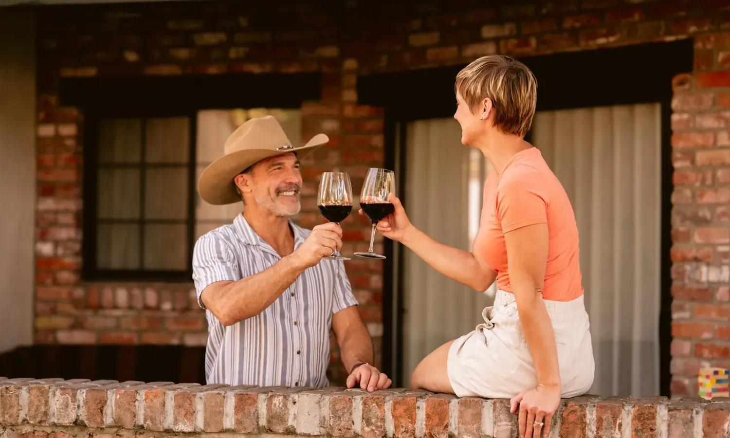 A man in a cowboy hat and a striped shirt drinking red wine with a woman in an orange top sitting on a brick wall.