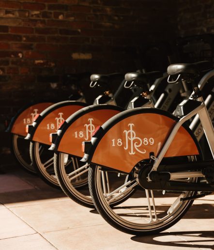 Four bikes with orange wheel covers displaying JRL 1889 are lined up against a brick wall in sunlight.