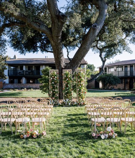 Rows of light wooden chairs are arranged on a grassy lawn facing a large tree, with three floral pillars decorated with greenery and colorful flowers at the front, set between two buildings.