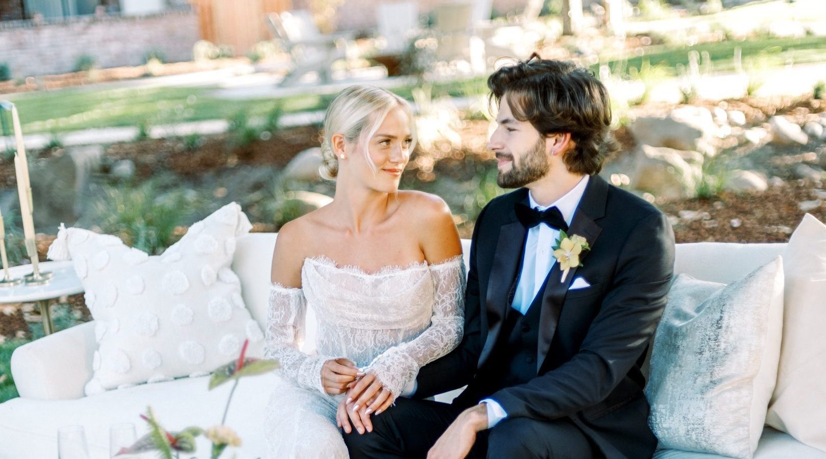 A bride in a white wedding dress sitting next the groom in a black suite on a white sofa in a garden.