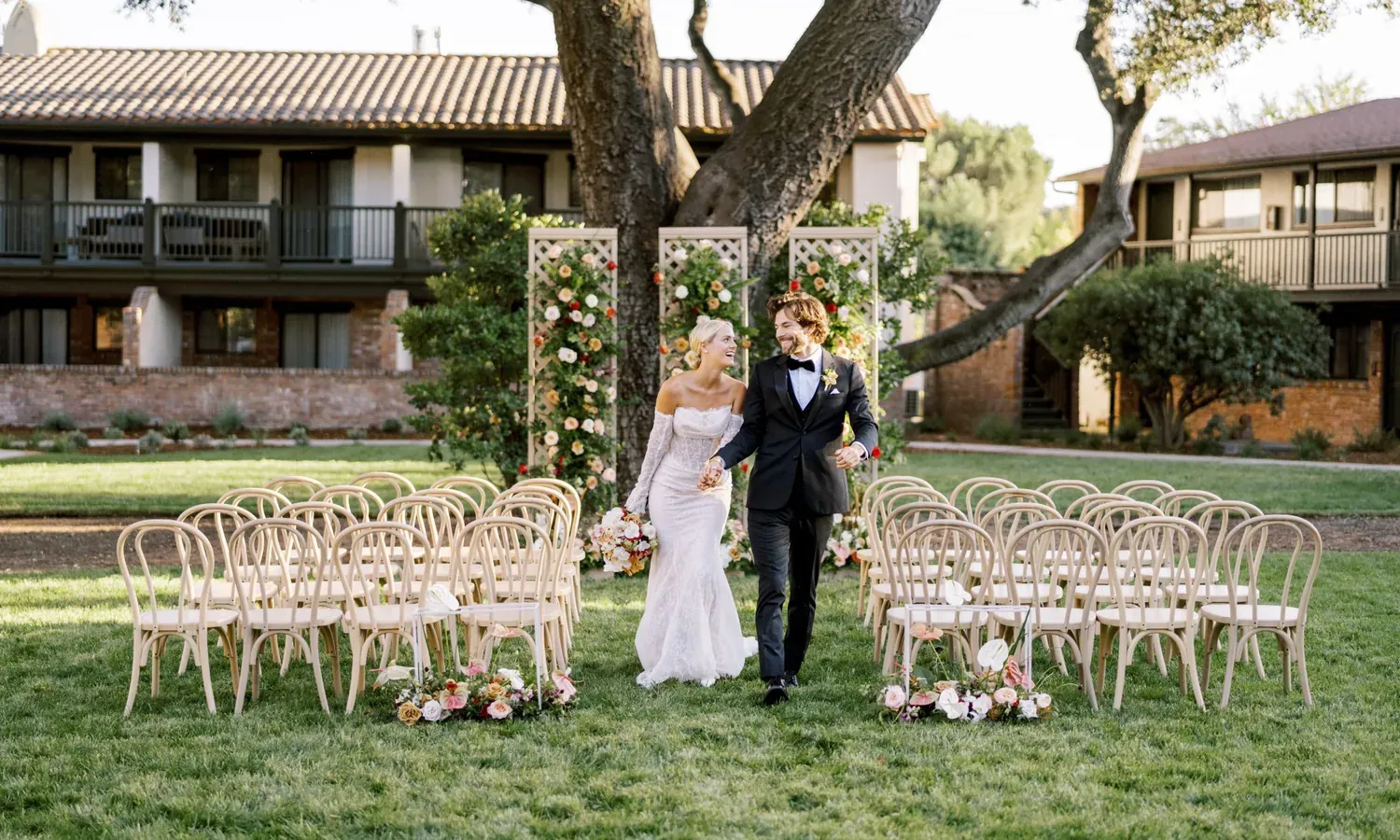 A bride in a white dress holding hands with the groom in a black suite running down the aisle of wooden wedding ceremony chairs in a garden.