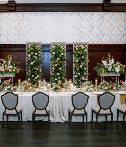An elegant table setting with gray chairs, a white tablecloth, floral centerpieces, tall candles, and three vertical trellises decorated with green foliage and colorful flowers against a patterned wall.