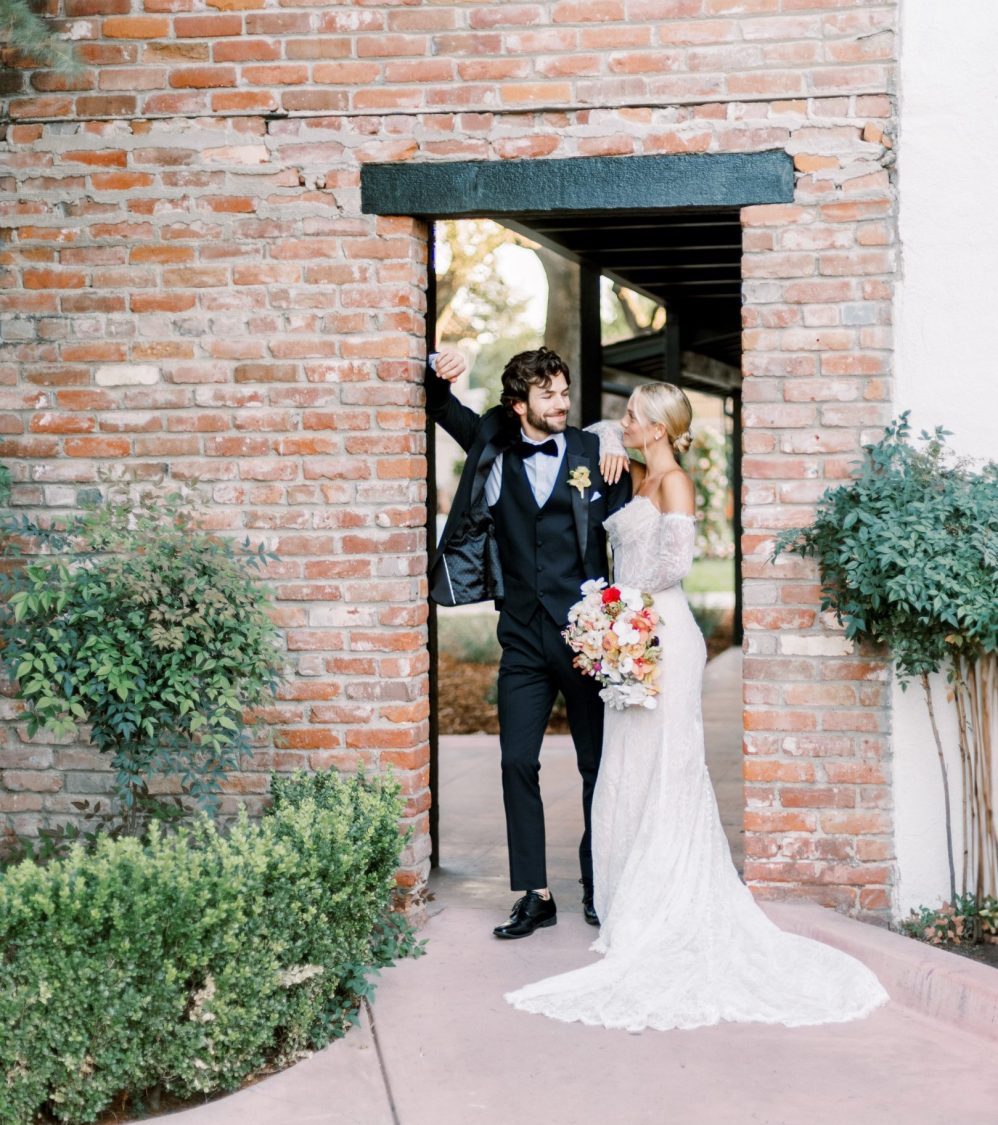 Groom in a black suite with bride in a white wedding dress leaning against a brick wall.