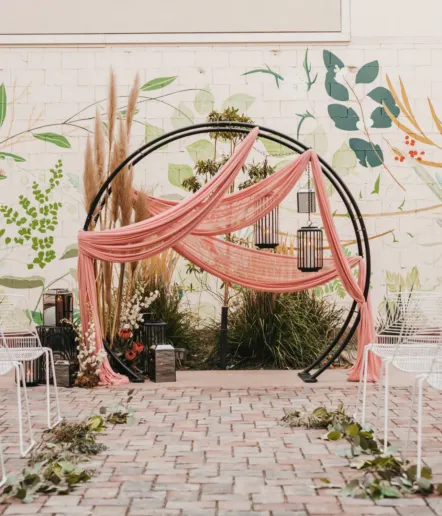 Outdoor wedding arch decorated with flowing pink fabric and black lanterns, surrounded by grasses and pampas, set on a brick patio with a leafy mural and white chairs arranged on either side.