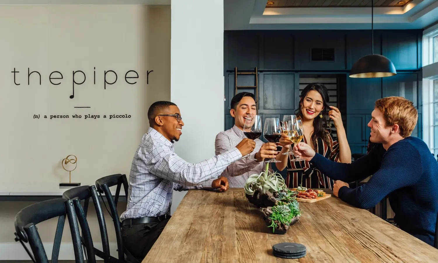 Four people sit around a wooden table in a modern kitchen, smiling and raising glasses in a toast. A sign on the wall reads the piper (n): a person who plays a piccolo. Food and plants are on the table.