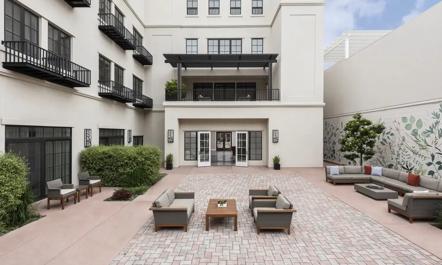 A spacious outdoor courtyard with brick flooring, patio sofas, chairs, and tables. The surrounding white building has balconies, large windows, and plants, with a mural of green leaves on the right wall.