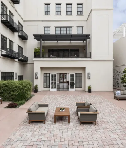 A spacious, modern courtyard with two sets of outdoor sofas and chairs around coffee tables on a paved patio, in front of a multi-story white building with balconies and large glass doors.