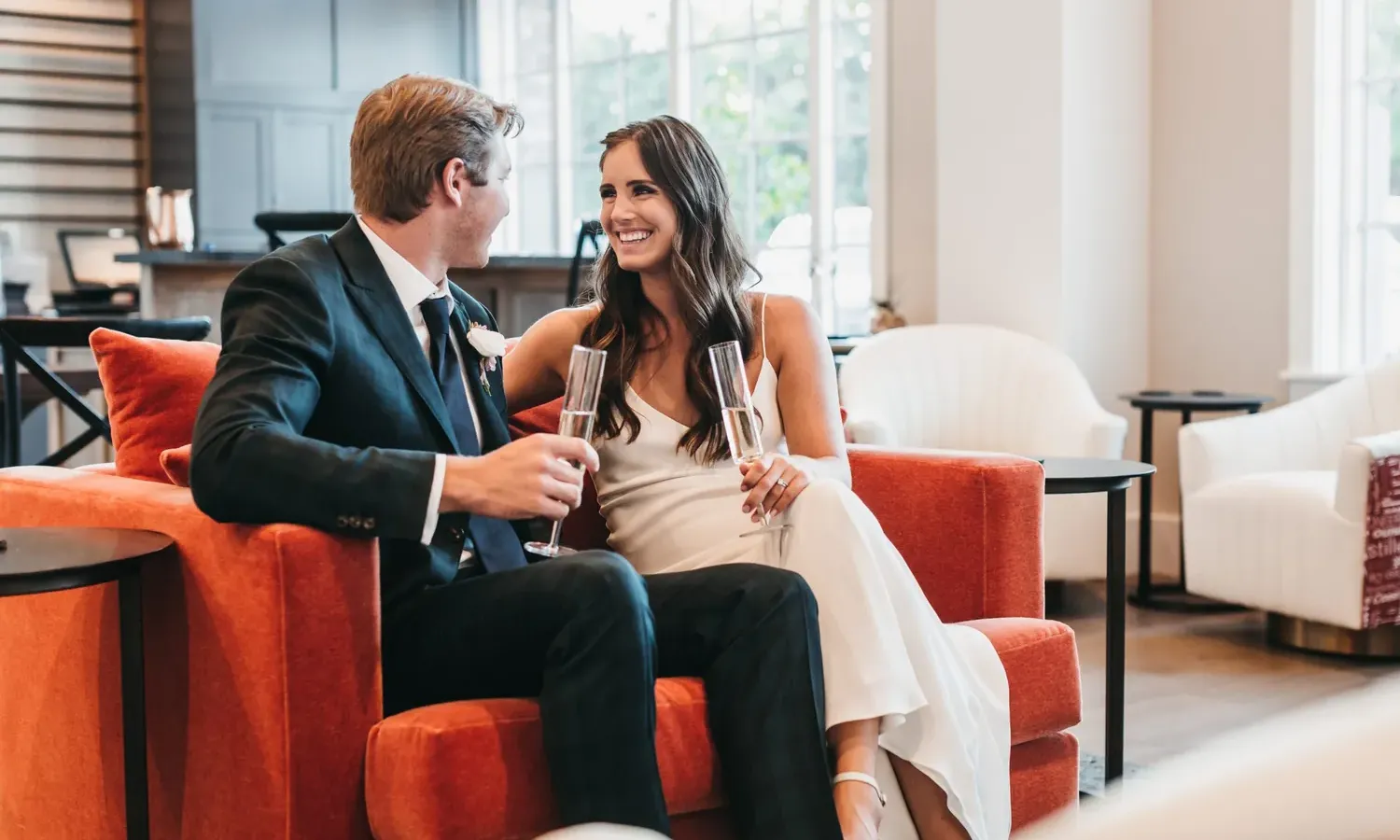 A man in a suit and a woman in a white dress sit close together on an orange couch, smiling at each other and holding champagne glasses in a bright, modern lounge.