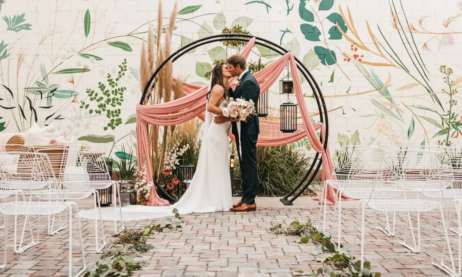 A bride and groom kiss in front of a circular wedding arch decorated with pink fabric and greenery, surrounded by white chairs and a wall mural featuring large botanical designs.