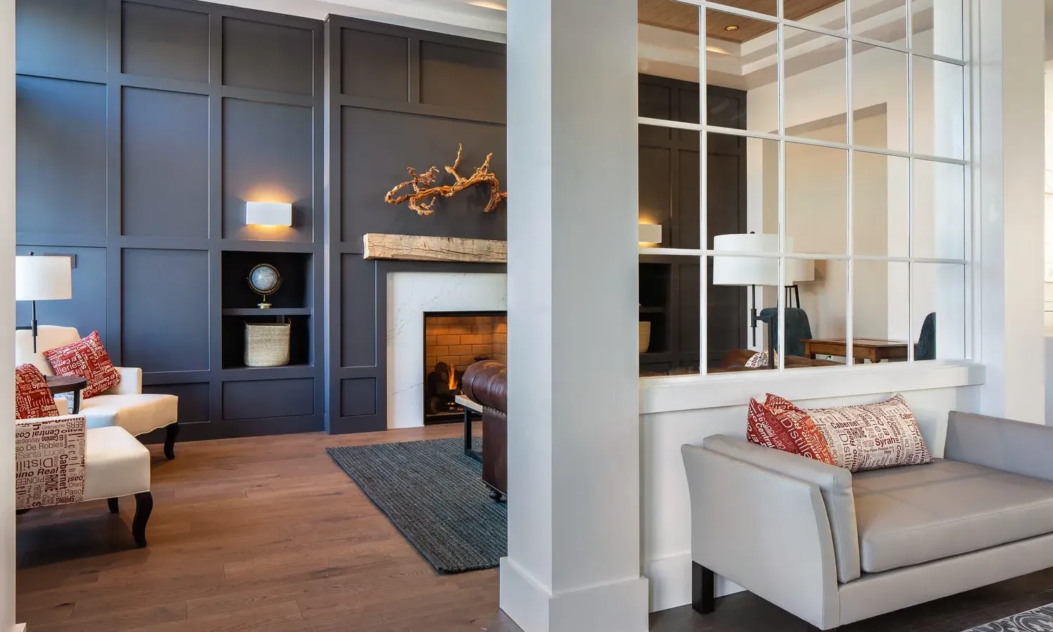 Modern living room with dark paneled accent wall, fireplace, white armchairs with red pillows, gray loveseat, hardwood floors, and large window with grid design separating spaces. Warm, cozy and elegantly decorated.