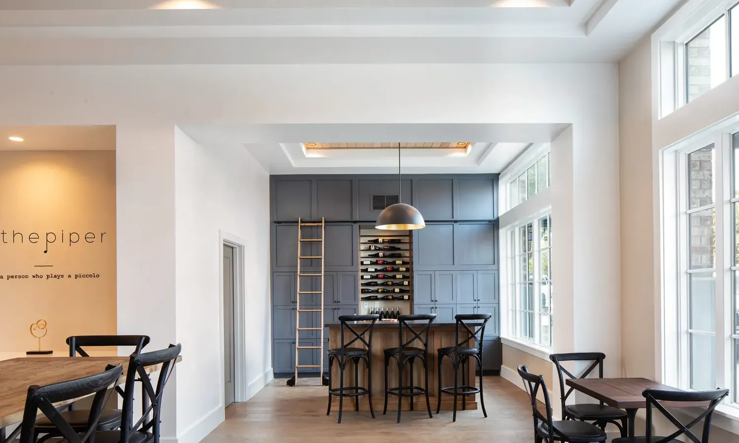 Modern dining area with wooden floors, black chairs around tables, a wall-mounted wine rack, tall gray cabinets, a metal ladder, and large windows letting in natural light. A black pendant lamp hangs above the central table.