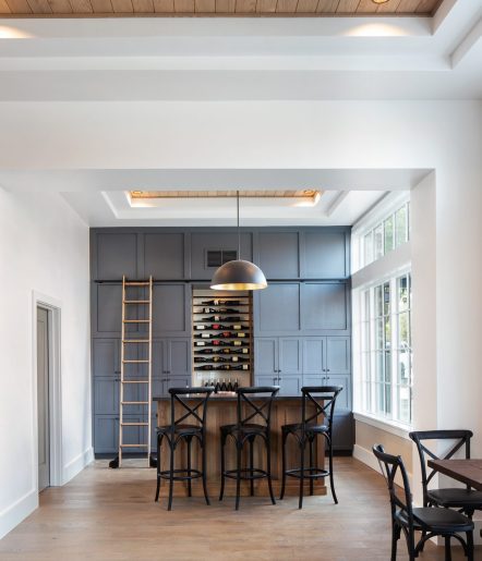 A modern home bar with a wooden countertop, black chairs, blue-gray paneled wall with built-in wine racks, a library ladder, and large windows allowing natural light into the room.