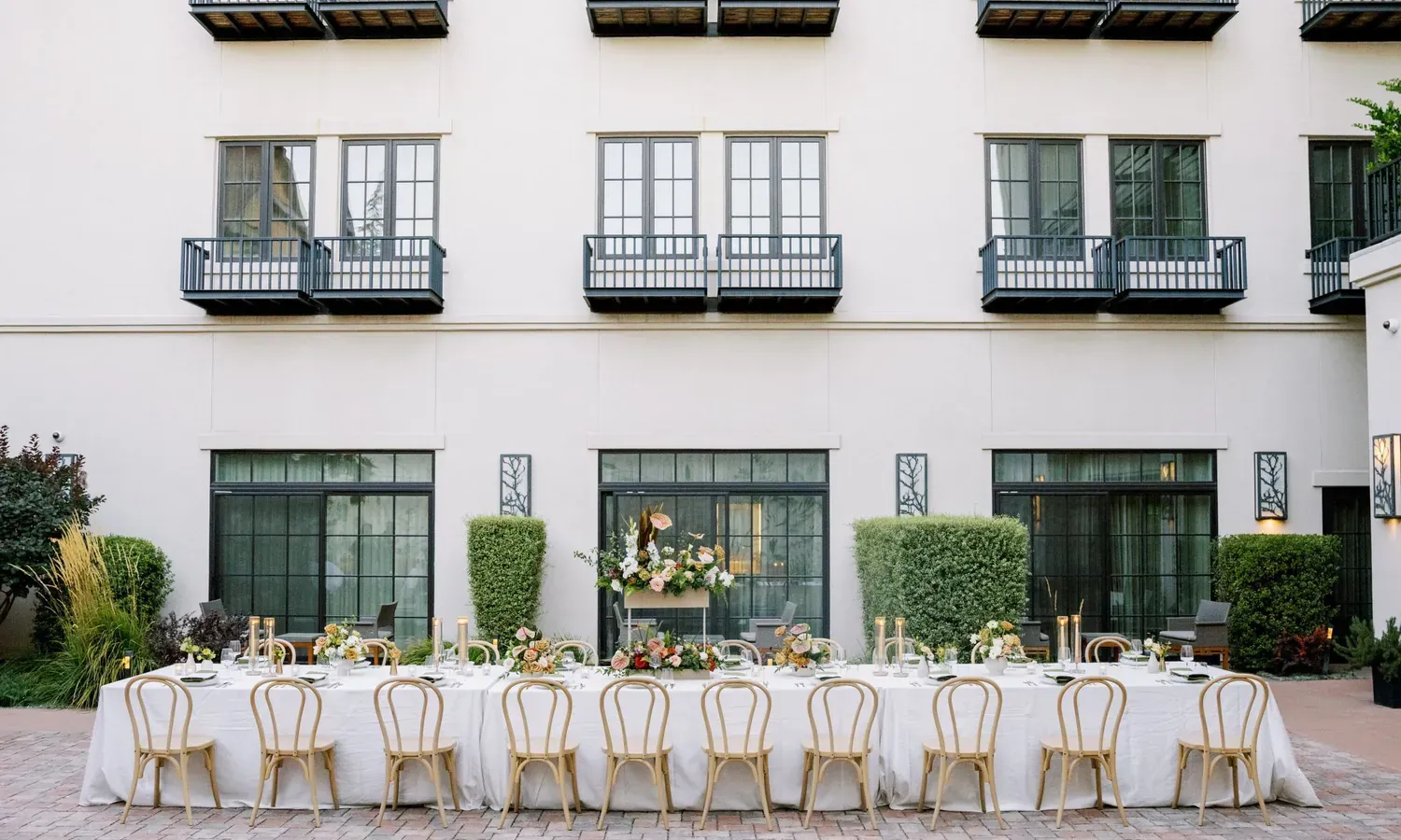 A long, elegant outdoor dining table with light wooden chairs, white tablecloth, and floral centerpieces is set in a courtyard in front of a modern building with large windows and small black balconies.