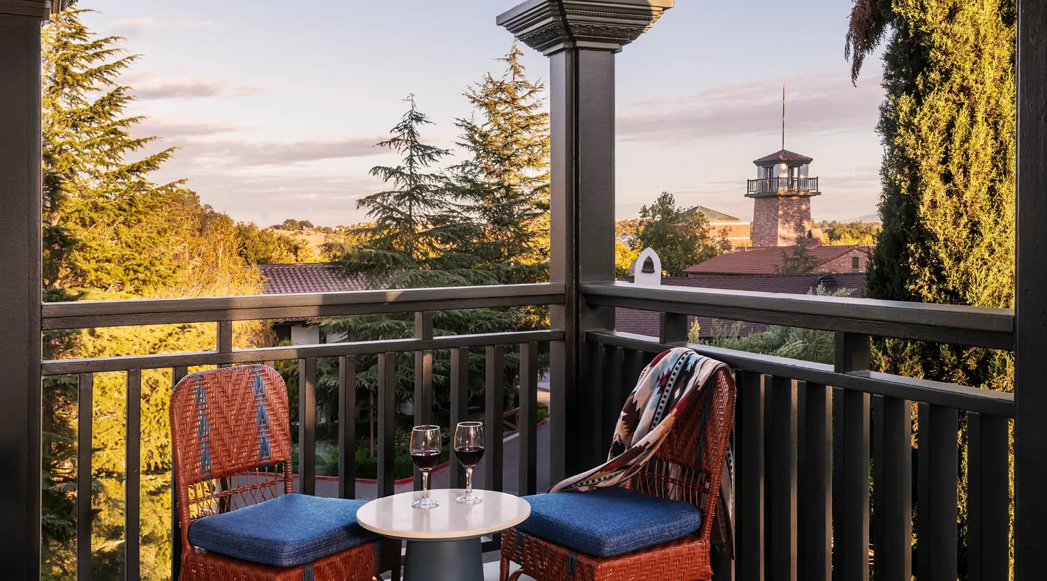 Two wicker chairs with blue cushions and a small table holding two glasses of red wine sit on a balcony overlooking trees and a building with a tower at sunset. A colorful blanket is draped over one chair.