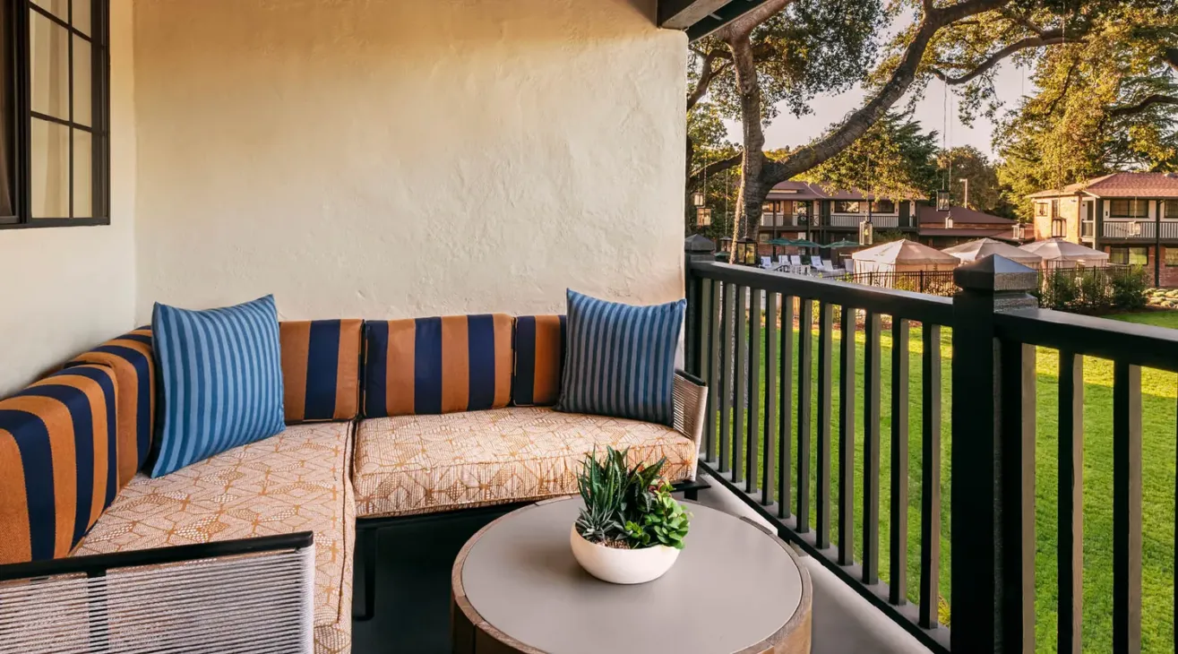 A cozy balcony with a striped L-shaped sofa, blue accent pillows, a round table with a potted plant, and a view of a green lawn, trees, and apartment buildings in the background.