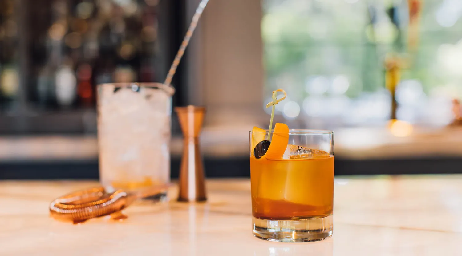 A glass of amber-colored cocktail with ice, garnished with an orange peel and cherry, sits on a marble bar. Behind it are a mixing glass with ice, a jigger, and a cocktail strainer, with a blurred bar background.