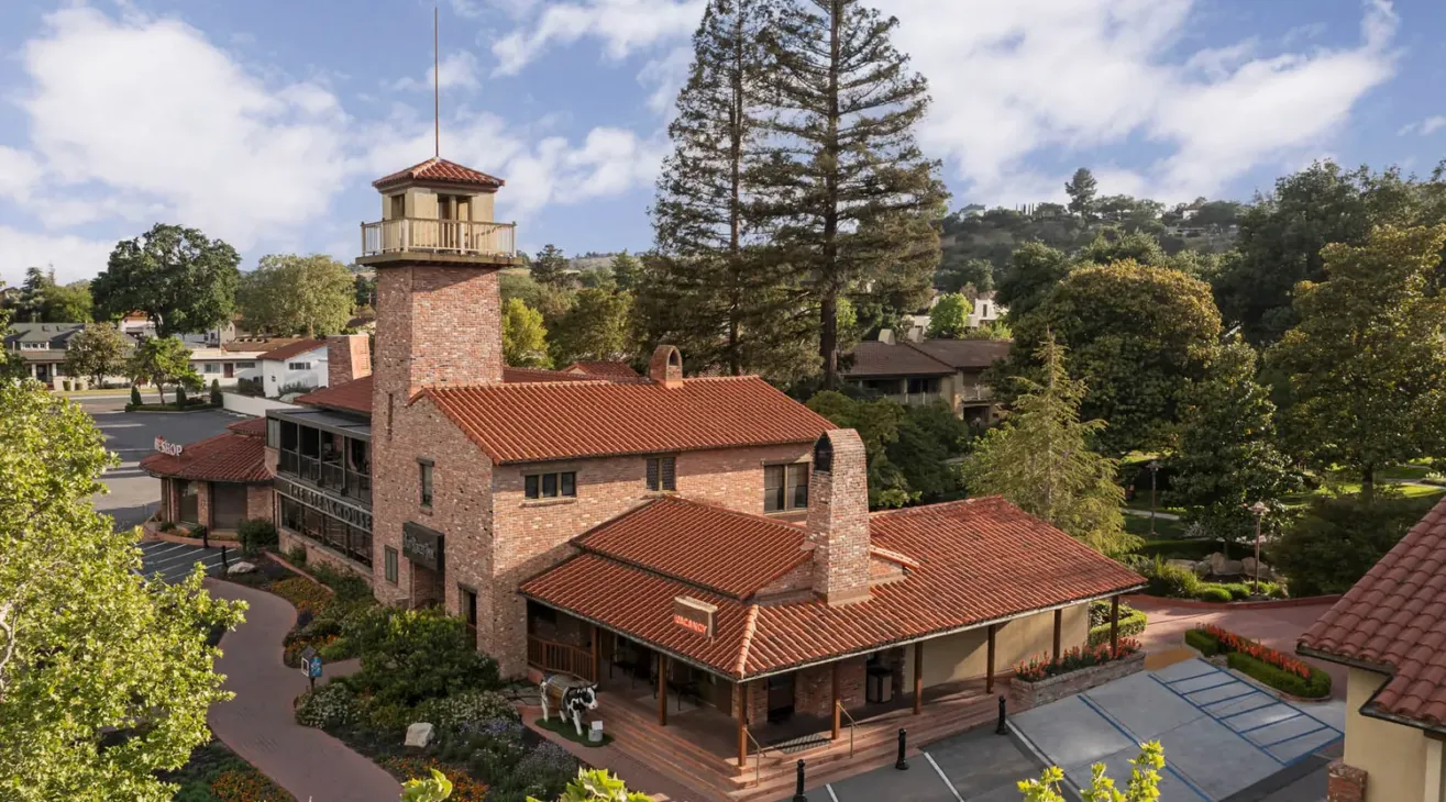 A brick building with a red tile roof and a tower, surrounded by trees and landscaping, sits in a suburban area under a partly cloudy sky. Accessible parking spaces are visible in front.