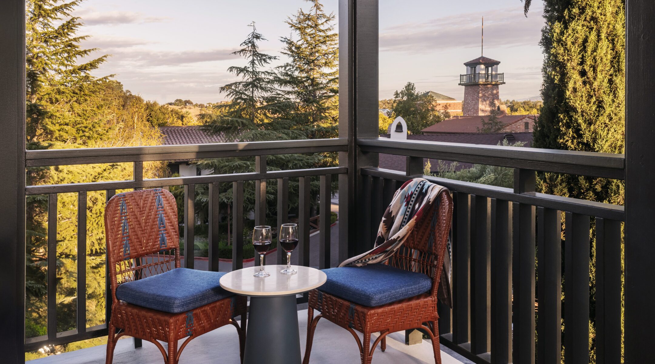 A cozy balcony at one of the hotels near Vina Robles Amphitheatre Paso Robles, featuring two wicker chairs, a small table with red wine, and a blanket. Enjoy sunset views of trees and a rustic building with a distant tower.