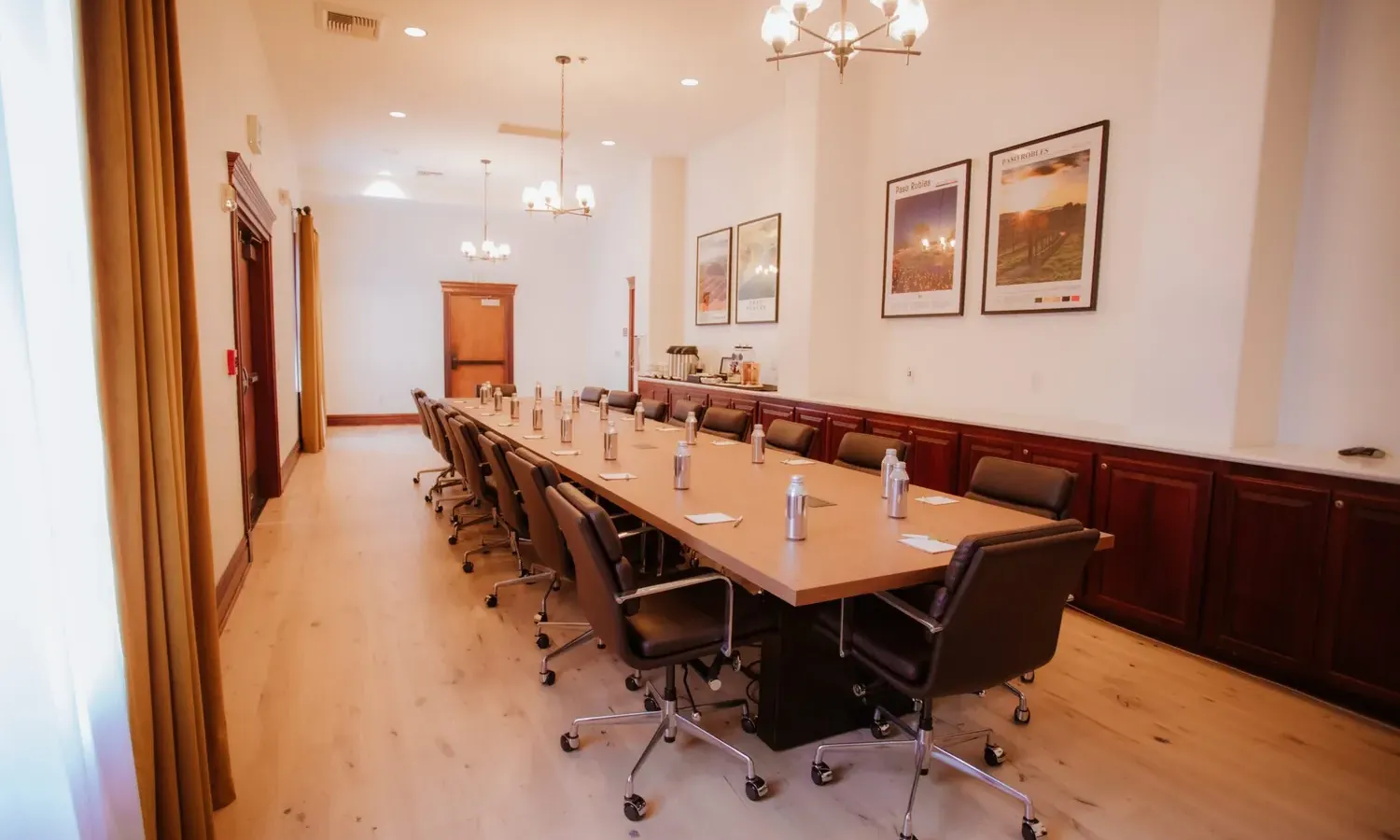 A modern conference room with a long wooden table surrounded by black chairs, water bottles placed at each seat, and framed posters on the white walls. The room is well-lit with chandeliers and natural light.