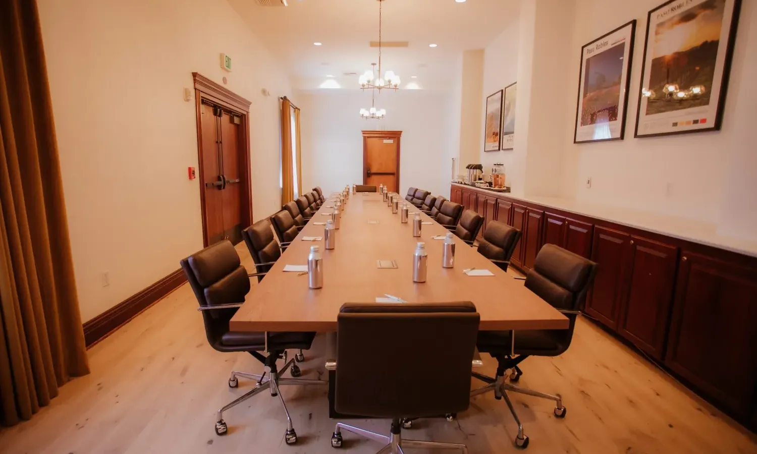 A modern conference room with a long wooden table, black office chairs, bottled water, notepads, and framed posters on the wall. The room has wood cabinets, chandeliers, and natural light from windows.