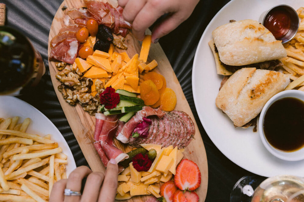 A top view of a charcuterie board with meats, cheeses, nuts, dried fruit, fresh strawberries, and cucumber slices—perfect for sharing after discovering local wine country hotel deals. Plates of fries and sandwiches surround the spread as hands reach for food.