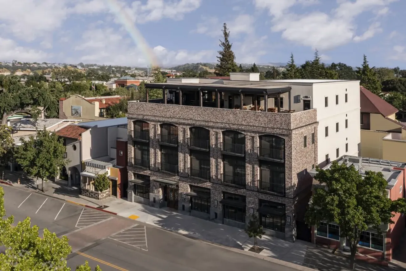 Aerial view of a boutique hotel in Paso Robles, housed in a modern brick building with large windows and a rooftop patio on a quiet street, surrounded by trees and neighboring buildings; a faint rainbow appears in the sky.