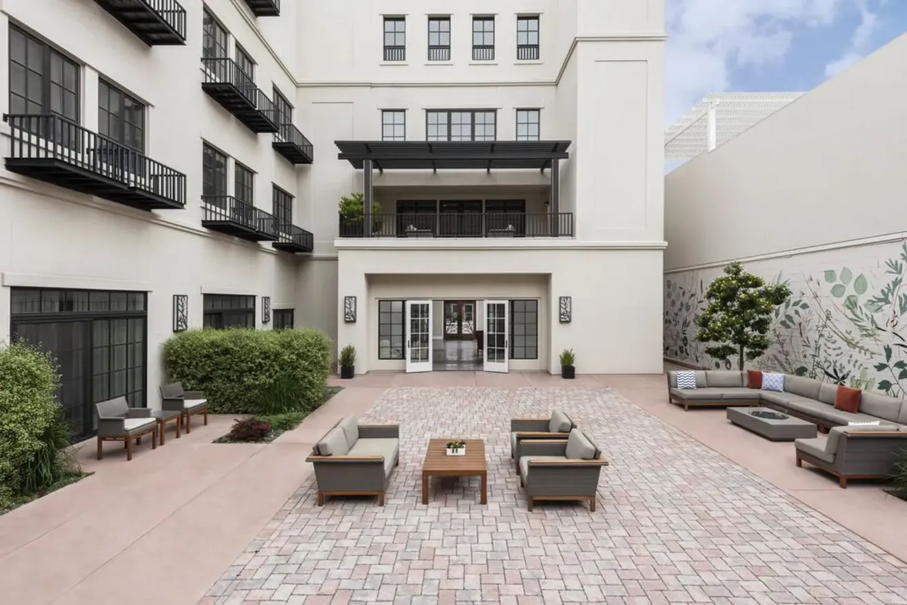A spacious outdoor patio at a boutique hotel in Paso Robles features cushioned seating, wooden tables, potted plants, and a mural, all surrounded by a multi-story building with balconies and large windows under a partly cloudy sky.