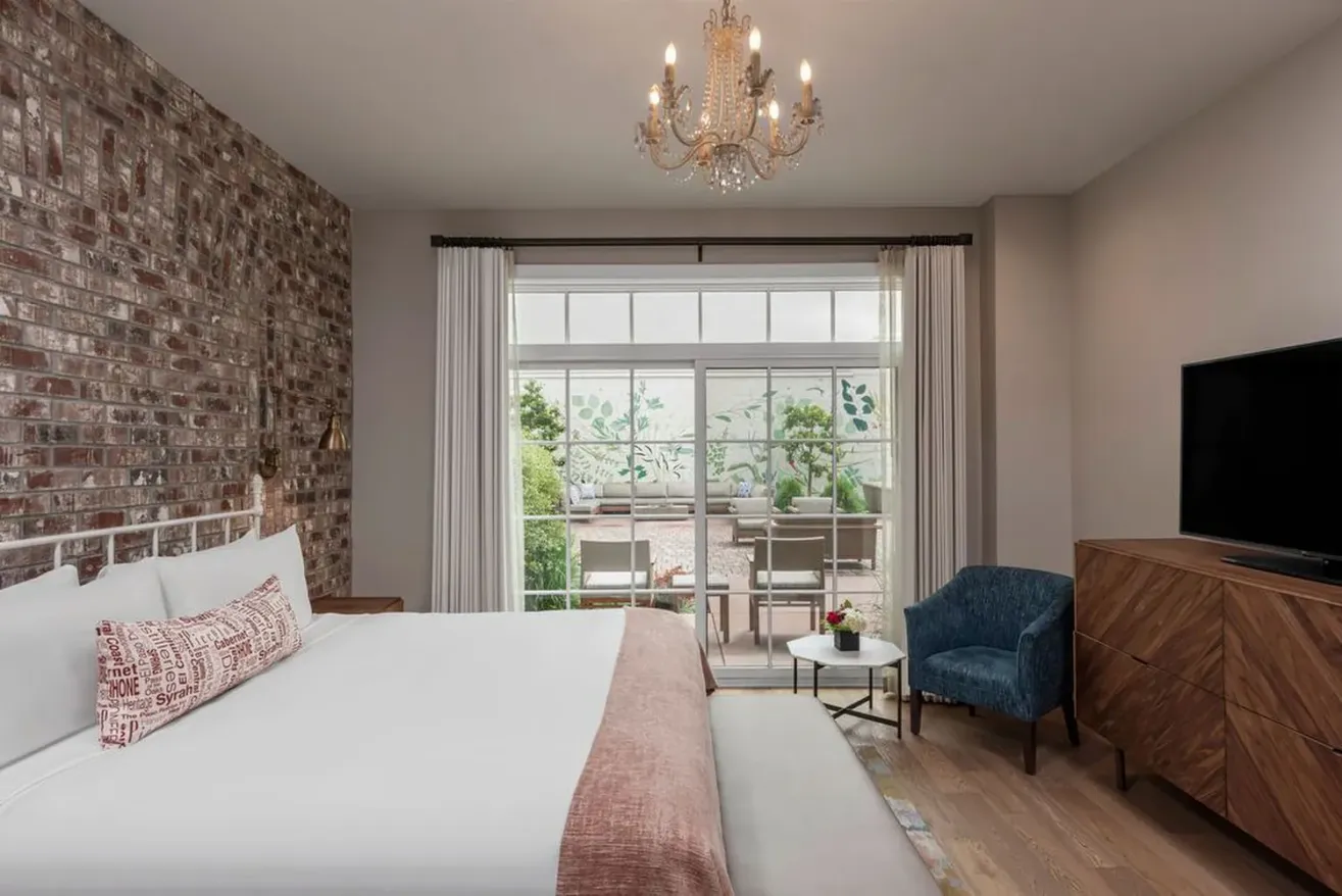 A modern boutique hotel in Paso Robles, featuring a brick accent wall, a white bed with pink accents, a chandelier, blue armchair, wooden dresser, TV, and large windows overlooking a patio with greenery.