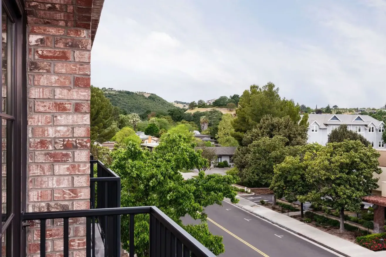 View from a brick balcony at a boutique hotel in Paso Robles, overlooking a quiet suburban street lined with trees and houses, with hills and greenery visible in the distance under a partly cloudy sky.
