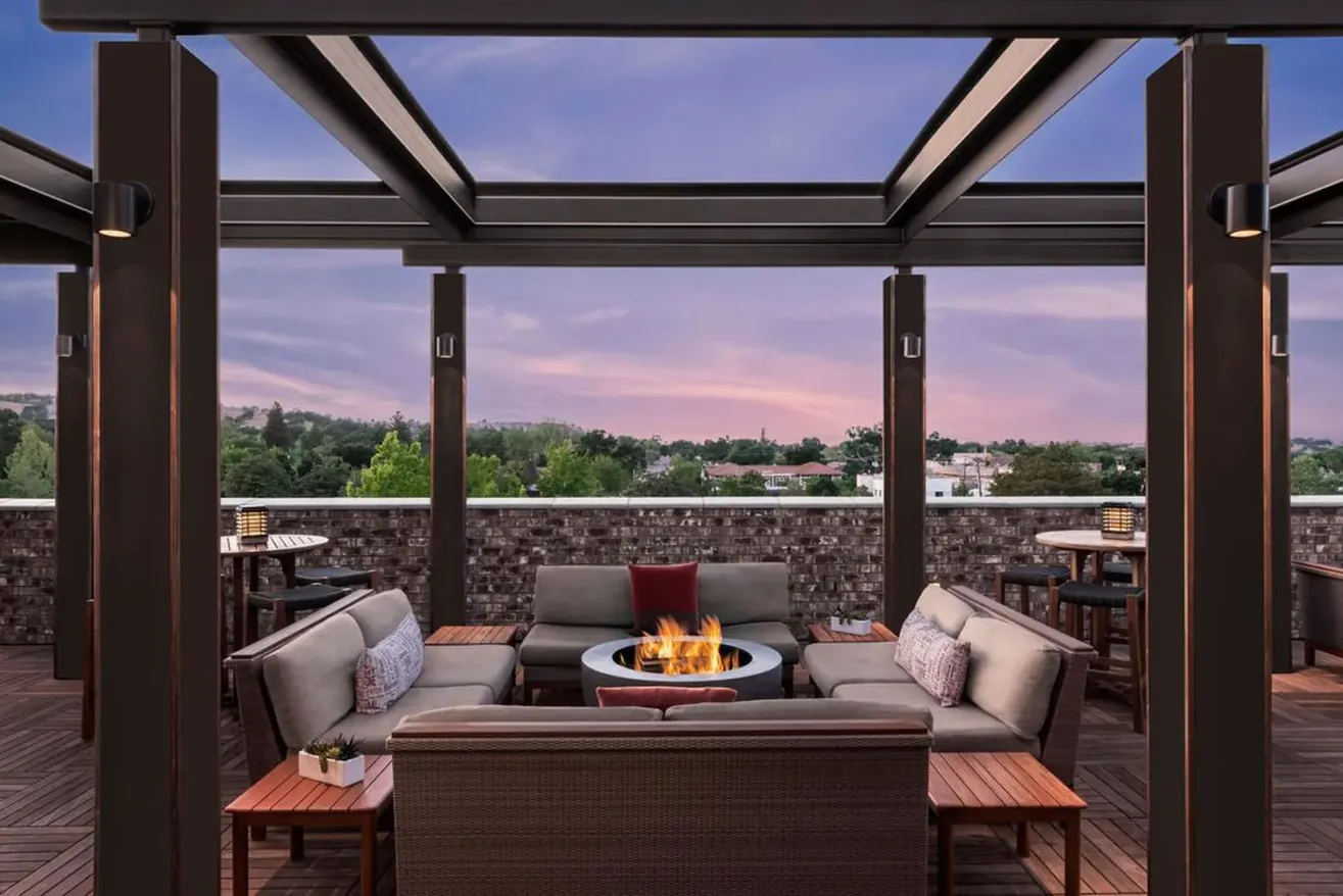Outdoor rooftop lounge at a boutique hotel in Paso Robles, featuring modern sofas around a fire pit, wooden tables, and an open pergola roof. Trees and buildings rise beyond the brick wall under a purple and pink evening sky.